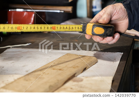 A carpenter holds a yellow tape measure above wooden board on a messy workshop desk, demonstrating precision and accuracy in building, home renovation, or manual crafting project tasks A carpenter holds a yellow tape measure above wooden board on a messy workshop desk, demonstrating precision and accuracy in building, home renovation, or manual crafting project tasks 142039770