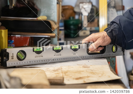 A senior builder holds a metallic spirit level over a wooden surface in a workshop, showcasing precise measurement and professional craftsmanship in home improvement projects A senior builder holds a metallic spirit level over a wooden surface in a workshop, showcasing precise measurement and professional craftsmanship in home improvement projects 142039786