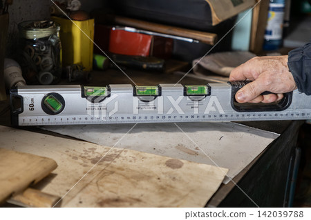 A hand holding a professional spirit level on a workbench, showing precision measurement tools during carpentry or construction work in a workshop to ensure flat surface accuracy A hand holding a professional spirit level on a workbench, showing precision measurement tools during carpentry or construction work in a workshop to ensure flat surface accuracy 142039788