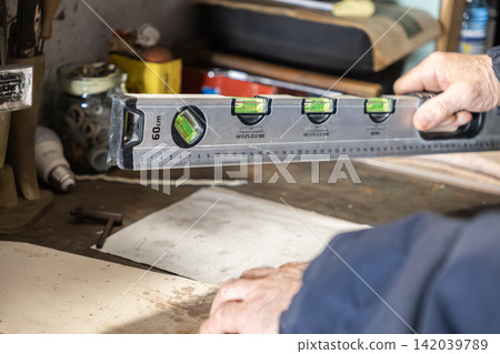 A senior artisan holds a silver spirit level tool over a wooden workbench in a workshop, indicating precision measurement during a construction project in a professional repair setting A senior artisan holds a silver spirit level tool over a wooden workbench in a workshop, indicating precision measurement during a construction project in a professional repair setting 142039789