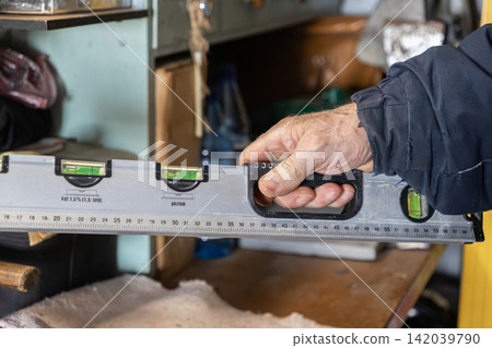 A male hand holds a grey spirit level tool in a workshop setting, symbolizing precision measurement, construction accuracy, professional maintenance, and skilled manual labor tasks A male hand holds a grey spirit level tool in a workshop setting, symbolizing precision measurement, construction accuracy, professional maintenance, and skilled manual labor tasks 142039790
