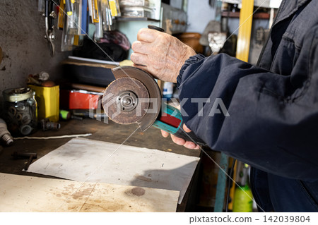 An elderly craftsman holds a power angle grinder with an abrasive disc in his workshop, embodying precision and skill in a professional manual task performed in a cluttered environment An elderly craftsman holds a power angle grinder with an abrasive disc in his workshop, embodying precision and skill in a professional manual task performed in a cluttered environment 142039804