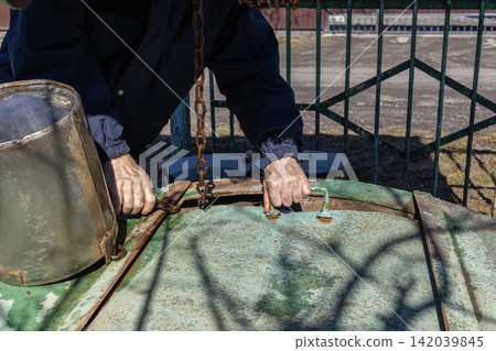 An elderly man pulls the heavy metal lid of a rustic water well using a handle, showcasing a daily rural chore involving traditional equipment in a quiet outdoor farm backyard setting An elderly man pulls the heavy metal lid of a rustic water well using a handle, showcasing a daily rural chore involving traditional equipment in a quiet outdoor farm backyard setting 142039845