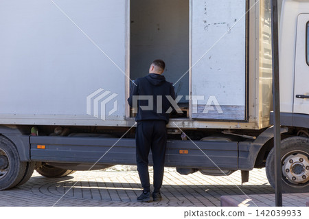 A delivery worker in a black hoodie loads parcels into a white truck, embodying efficiency and logistics services within a professional urban transportation supply chain context A delivery worker in a black hoodie loads parcels into a white truck, embodying efficiency and logistics services within a professional urban transportation supply chain context 142039933