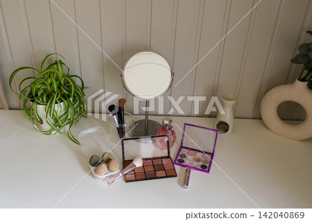 Vanity round mirror and make up items arranged on desk under lamp, palettes and brushes, open eyeshadow, cotton pads. Makeup and self care at home concept. Personal beauty products items on table. Vanity round mirror and make up items arranged on desk under lamp, palettes and brushes, open eyeshadow, cotton pads. Makeup and self care at home concept. Personal beauty products items on table. 142040869