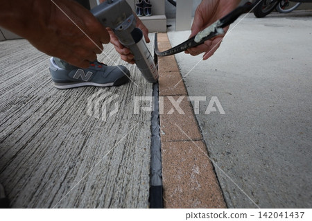 Two workers are seen applying caulking to the expansion joint between concrete and brick. Two workers are seen applying caulking to the expansion joint between concrete and brick. 142041437