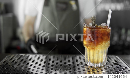 Refreshing layered cocktail with ice and rosemary on bar counter, bartender working in background Refreshing layered cocktail with ice and rosemary on bar counter, bartender working in background 142041664