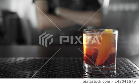 Negroni cocktail with a vibrant orange peel on a bar counter, bartender in blurred monochrome background Negroni cocktail with a vibrant orange peel on a bar counter, bartender in blurred monochrome background 142041679