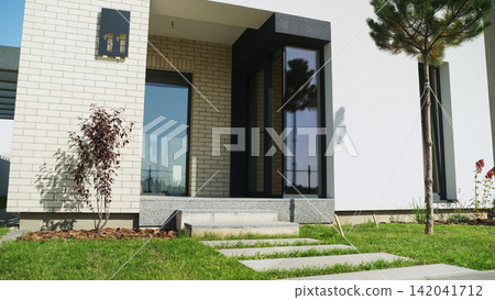 Modern house entrance featuring clean lines, large glass door, white and brick walls, concrete pathway, and green lawn Modern house entrance featuring clean lines, large glass door, white and brick walls, concrete pathway, and green lawn 142041712