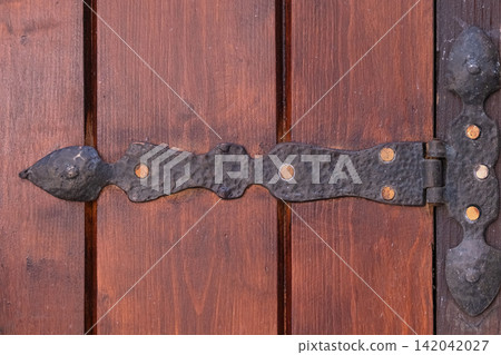 Ornate dark metal hinge securing a weathered brown wooden door, showcasing handcrafted hardware and traditional craftsmanship, reflecting history and vintage style Ornate dark metal hinge securing a weathered brown wooden door, showcasing handcrafted hardware and traditional craftsmanship, reflecting history and vintage style 142042027
