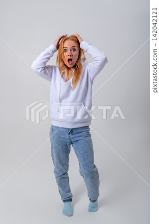 Young woman with red hair expressing shock and disbelief while holding her head with both hands, mouth open and eyes wide, standing on a light background Young woman with red hair expressing shock and disbelief while holding her head with both hands, mouth open and eyes wide, standing on a light background 142042121