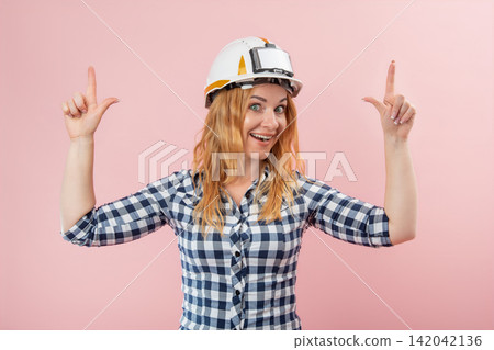 Happy young woman wearing a construction hard hat and plaid shirt, enthusiastically pointing with both index fingers upwards, presenting a new idea or important information Happy young woman wearing a construction hard hat and plaid shirt, enthusiastically pointing with both index fingers upwards, presenting a new idea or important information 142042136