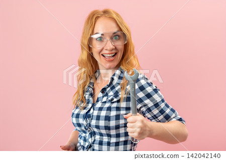 Young woman with red hair wearing a plaid shirt and protective glasses, enthusiastically holding a wrench on a pink background, representing female empowerment in skilled trades Young woman with red hair wearing a plaid shirt and protective glasses, enthusiastically holding a wrench on a pink background, representing female empowerment in skilled trades 142042140