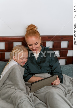 Mother and daughter lying in bed under a cozy blanket, sharing a happy moment while watching content on a digital tablet, illustrating family bonding and leisure Mother and daughter lying in bed under a cozy blanket, sharing a happy moment while watching content on a digital tablet, illustrating family bonding and leisure 142042159