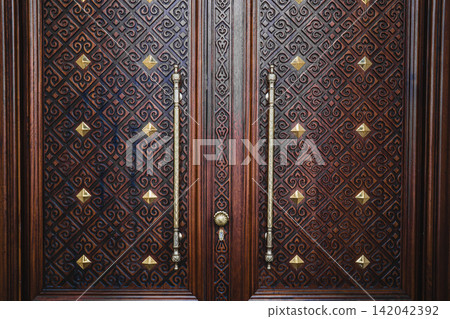 Ornate carved dark wooden double door with golden brass studs in mosque, traditional Islamic architecture concept Ornate carved dark wooden double door with golden brass studs in mosque, traditional Islamic architecture concept 142042392