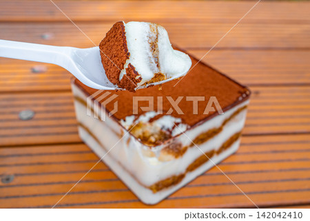 Spoon lifting slice of tiramisu dessert with cream and cocoa layers on wooden table, Italian food concept Spoon lifting slice of tiramisu dessert with cream and cocoa layers on wooden table, Italian food concept 142042410