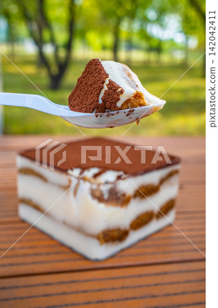 Spoon lifting slice of tiramisu dessert with cream and cocoa layers on wooden table, Italian food concept Spoon lifting slice of tiramisu dessert with cream and cocoa layers on wooden table, Italian food concept 142042411