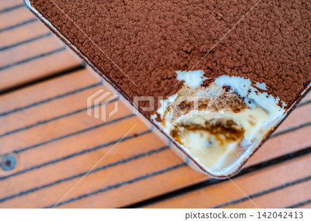Close up of tiramisu dessert in white tray showing cream layers on wooden surface, Italian food concept Close up of tiramisu dessert in white tray showing cream layers on wooden surface, Italian food concept 142042413