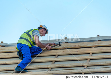 Construction worker wearing a hard hat and safety vest installing horizontal wooden battens on a new roof framing with a hammer, preparing for weatherproofing and tile placement Construction worker wearing a hard hat and safety vest installing horizontal wooden battens on a new roof framing with a hammer, preparing for weatherproofing and tile placement 142042421