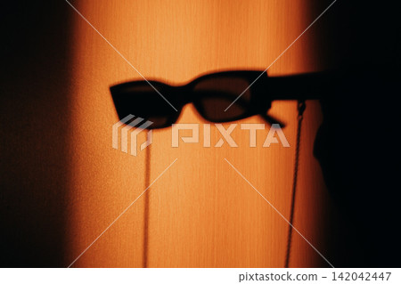 Black sunglasses casting a dramatic shadow on a warm orange wall, creating a bold minimalist image with strong contrast, mystery, fashion, summer mood, and contemporary visual style and graphic form. Black sunglasses casting a dramatic shadow on a warm orange wall, creating a bold minimalist image with strong contrast, mystery, fashion, summer mood, and contemporary visual style and graphic form. 142042447