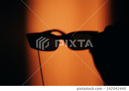 Black sunglasses casting a dramatic shadow on a warm orange wall, creating a bold minimalist image with strong contrast, mystery, fashion, summer mood, and contemporary visual style and graphic form. Black sunglasses casting a dramatic shadow on a warm orange wall, creating a bold minimalist image with strong contrast, mystery, fashion, summer mood, and contemporary visual style and graphic form. 142042448