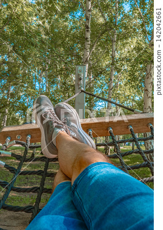 Person resting feet on wooden park net hammock swing in birch tree summer park, outdoor leisure lifestyle concept 142042661