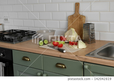 Simple home cooking. Wooden table with fresh vegetables, sliced cabbage, halved tomatoes, cucumber, cutting board knife. Preparing nutritious midday meals. Creating nourishing. Assembling hearty lunch 142042819