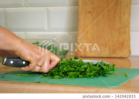 Person Chopping parsley bunch. Female cut greens with sharp knife at home kitchen. Woman hands cutting fresh aromatic herbs for cooking. Preparing food. Healthy food ingredient. Ecological products. Person Chopping parsley bunch. Female cut greens with sharp knife at home kitchen. Woman hands cutting fresh aromatic herbs for cooking. Preparing food. Healthy food ingredient. Ecological products. 142042833