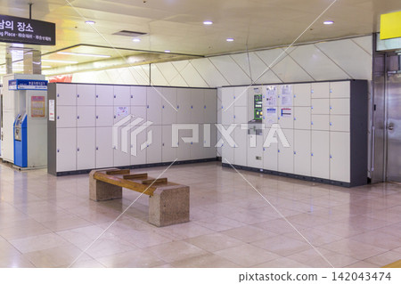 An empty public transit station interior features rows of storage lockers, wooden benches, tiled flooring, ceiling lights, and directional signs with Korean text. An empty public transit station interior features rows of storage lockers, wooden benches, tiled flooring, ceiling lights, and directional signs with Korean text. 142043474