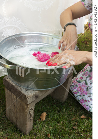 A rustic scene featuring a person in a floral dress holding a handmade soap bar wrapped in white cloth over a metal bucket. A rustic scene featuring a person in a floral dress holding a handmade soap bar wrapped in white cloth over a metal bucket. 142043927