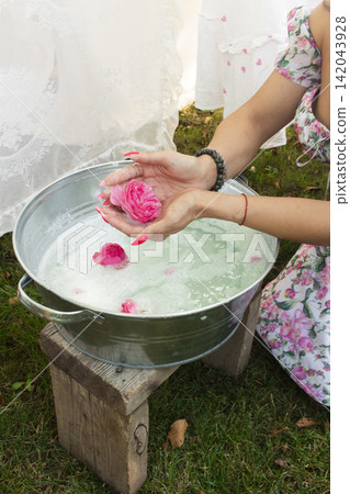 A rustic scene featuring a person in a floral dress holding a handmade soap bar wrapped in white cloth over a metal bucket. A rustic scene featuring a person in a floral dress holding a handmade soap bar wrapped in white cloth over a metal bucket. 142043928