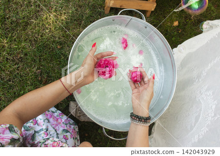 A rustic scene featuring a person in a floral dress holding a handmade soap bar wrapped in white cloth over a metal bucket. A rustic scene featuring a person in a floral dress holding a handmade soap bar wrapped in white cloth over a metal bucket. 142043929