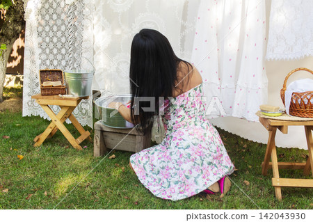 A rustic outdoor laundry scene featuring a woman in a floral dress kneeling on grass, washing clothes by hand in a metal basin placed on a wooden stool. A rustic outdoor laundry scene featuring a woman in a floral dress kneeling on grass, washing clothes by hand in a metal basin placed on a wooden stool. 142043930