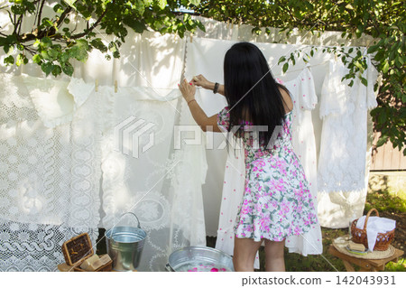 A rustic outdoor laundry scene featuring a woman in a floral dress kneeling on grass, washing clothes by hand in a metal basin placed on a wooden stool. A rustic outdoor laundry scene featuring a woman in a floral dress kneeling on grass, washing clothes by hand in a metal basin placed on a wooden stool. 142043931