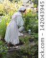 A serene outdoor gardening scene featuring a person in a light dress with a white apron and shoes, watering plants with a metal watering can. 142043945
