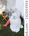 A rustic outdoor laundry scene featuring a woman in a pastel dress with ruffled sleeves, apron, and lace head covering, kneeling on grass. 142043954