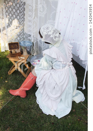A rustic outdoor laundry scene featuring a woman in a pastel dress with ruffled sleeves, apron, and lace head covering, kneeling on grass. A rustic outdoor laundry scene featuring a woman in a pastel dress with ruffled sleeves, apron, and lace head covering, kneeling on grass. 142043954