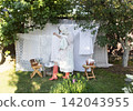 A rustic outdoor laundry scene featuring a woman in a pastel dress with ruffled sleeves, apron, and lace head covering, kneeling on grass. 142043955