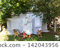 A rustic outdoor laundry scene featuring a woman in a pastel dress with ruffled sleeves, apron, and lace head covering, kneeling on grass. 142043956