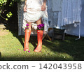 A rustic outdoor scene featuring a person in a light dress with lace trim and bright orange rain boots used as flower pots, filled with purple blossoms. 142043958