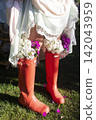 A rustic outdoor scene featuring a person in a light dress with lace trim and bright orange rain boots used as flower pots, filled with purple blossoms. 142043959