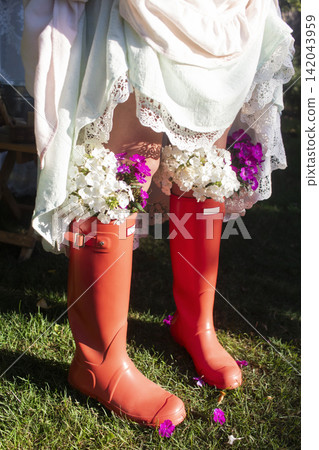 A rustic outdoor scene featuring a person in a light dress with lace trim and bright orange rain boots used as flower pots, filled with purple blossoms. A rustic outdoor scene featuring a person in a light dress with lace trim and bright orange rain boots used as flower pots, filled with purple blossoms. 142043959