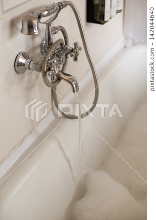 Water running from vintage chrome faucet into bathtub with foam in bathroom 142044640