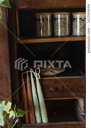 Handmade wax taper candles and dried herbs jars in vintage wooden cabinet Handmade wax taper candles and dried herbs jars in vintage wooden cabinet 142044644