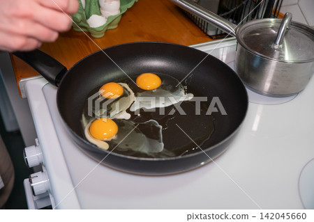 Hand cooks three sunny side up eggs in black frying pan on electric stovetop. Breakfast preparation, home cooking, homemade meal, simple nutrition, kitchen routine, everyday food. Hand cooks three sunny side up eggs in black frying pan on electric stovetop. Breakfast preparation, home cooking, homemade meal, simple nutrition, kitchen routine, everyday food. 142045660