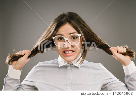 Woman, portrait and pulling hair in studio with stress for education, exam or assessment by background. Girl, college student or frustrated person with anxiety for study, test or glasses for learning Woman, portrait and pulling hair in studio with stress for education, exam or assessment by background. Girl, college student or frustrated person with anxiety for study, test or glasses for learning 142045908