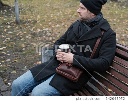 Pensive hipster drinking coffee in the urban street. Handsome man wearing black coat, brown leather bag walking confidently down the city with paper cup in hand. Stylish bearded middle-aged male. Pensive hipster drinking coffee in the urban street. Handsome man wearing black coat, brown leather bag walking confidently down the city with paper cup in hand. Stylish bearded middle-aged male. 142045952