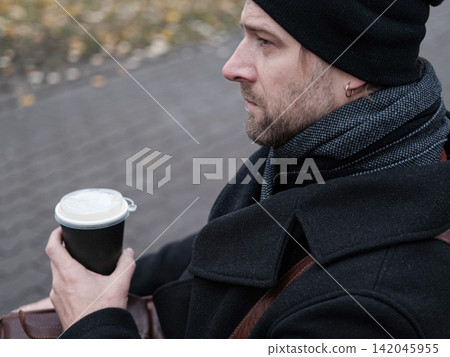 Pensive hipster drinking coffee in the urban street. Handsome man wearing black coat, brown leather bag walking confidently down the city with paper cup in hand. Stylish bearded middle-aged male. Pensive hipster drinking coffee in the urban street. Handsome man wearing black coat, brown leather bag walking confidently down the city with paper cup in hand. Stylish bearded middle-aged male. 142045955