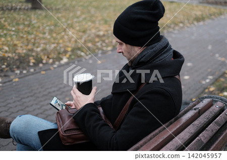 Pensive hipster drinking coffee in the urban street. Handsome man wearing black coat, brown leather bag walking confidently down the city with paper cup in hand. Stylish bearded middle-aged male. 142045957
