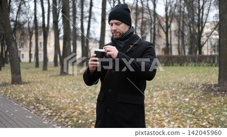 Pensive hipster drinking coffee in the urban street. Handsome man wearing black coat, brown leather bag walking confidently down the city with paper cup in hand. Stylish bearded middle-aged male. 142045960